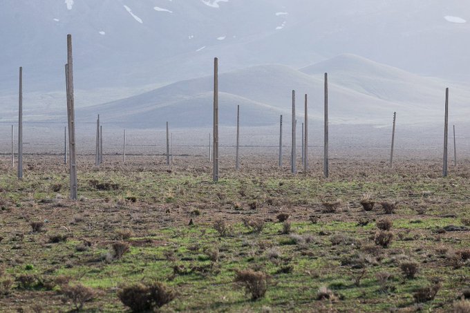 A barren landscape with patchy green and brown vegetation, scattered wooden utility poles, and snow-capped mountains in the distance.