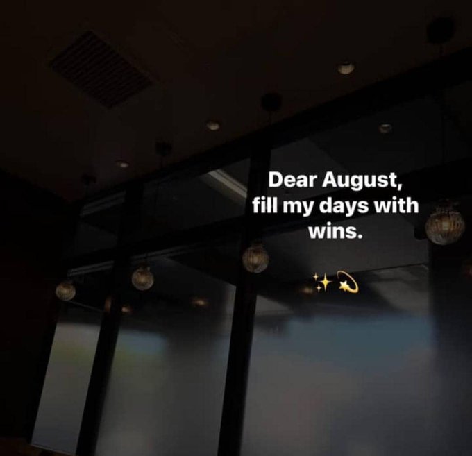 A dimly lit room with hanging pendant lights displays the white text 'Dear August, fill my days with wins.' accompanied by small star and Saturn emojis.