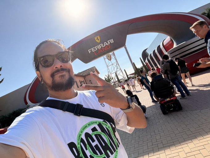 A man wearing sunglasses and a "Pasta" t-shirt points at the camera in a selfie taken at the entrance of Ferrari Land, a theme park, under a bright sun with other visitors visible in the background. The "FERRARI LAND" signage is prominently displayed on the arched entrance, suggesting a fun midweek outing at the amusement park.