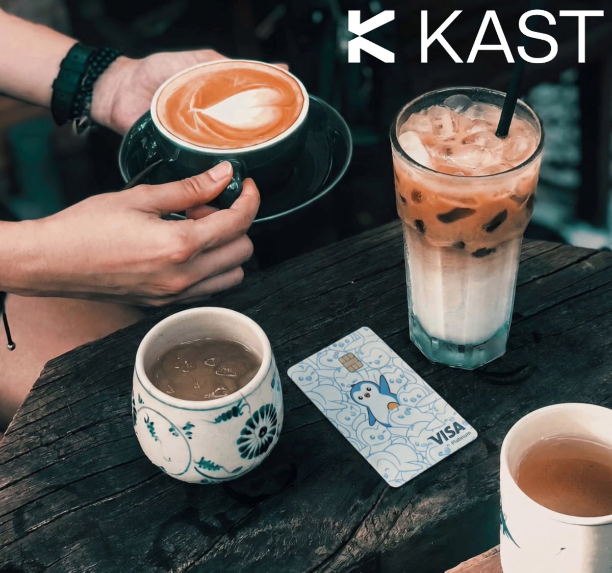 The image shows a person sitting at a table with two cups of coffee - one black cup and one white cup filled with lattes. There are also three other cups placed around the table, suggesting that this could be a gathering or social event. The person is holding a wallet in their hand, which appears to be a Visa card. The image promotes the use of @KAST for daily transactions, emphasizing the convenience and modernity it offers compared to traditional banking methods.