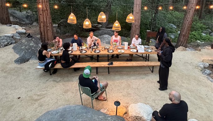 A group of people are gathered around a large wooden dining table set in a forested outdoor area with string lights and hanging lamps, as one person stands addressing the seated guests in what appears to be a casual dinner gathering related to the FWB FEST 2026 event.