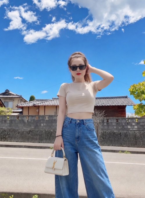 A woman wearing sunglasses, a beige crop top, and blue jeans poses outdoors on a sunny day holding a white handbag, with traditional Japanese-style buildings visible in the background.