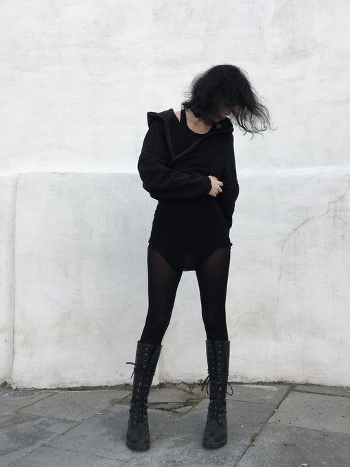 A person with long dark hair standing against a textured wall, wearing an oversized black hoodie, black tights, and tall lace-up black boots.