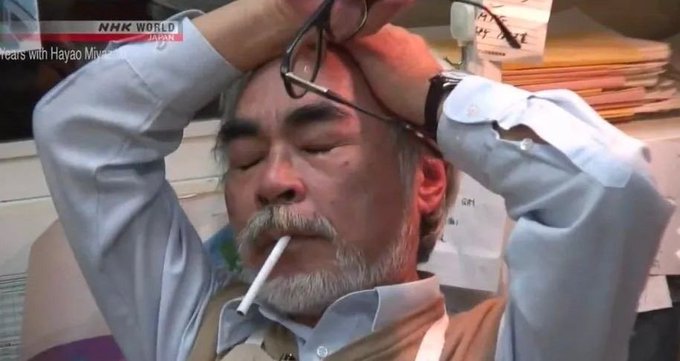 An older man with a gray beard smokes a cigarette while holding his glasses on top of his head, shown on an NHK World broadcast.
