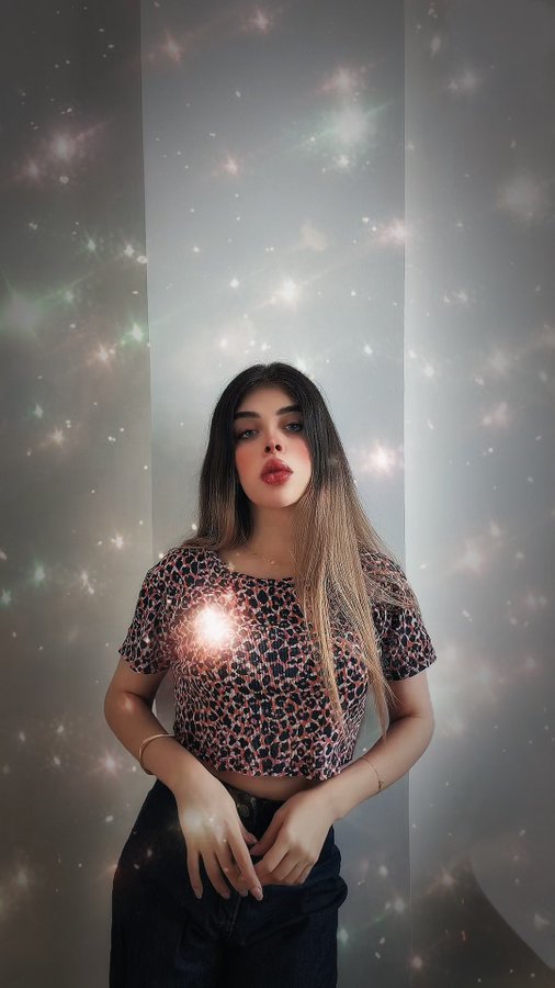 A selfie of a young woman with long dark hair wearing a leopard print top, set against a sparkling, galaxy-like background effect.