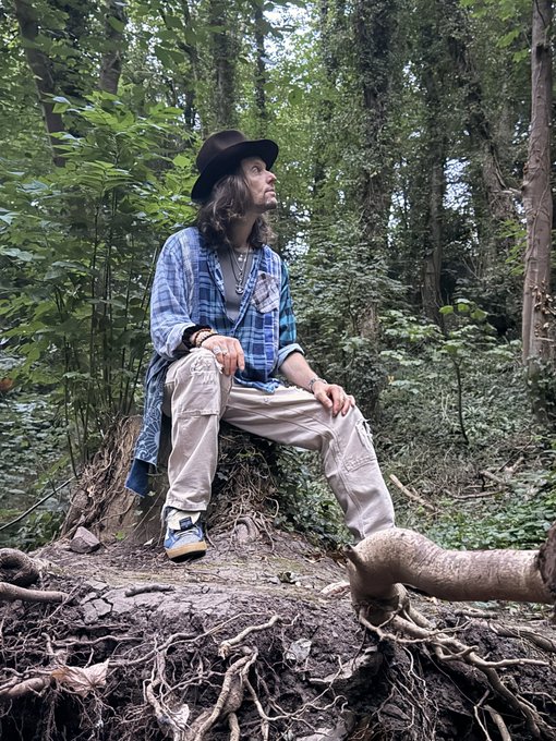 A man wearing a black hat, blue plaid shirt, and white pants sitting on a tree stump in a lush green forest, looking off to the side.