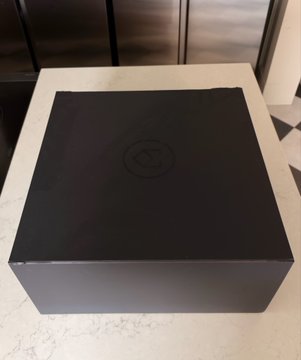 A large black box with a faint circular logo on top sits on a marble countertop in front of a stainless steel refrigerator.