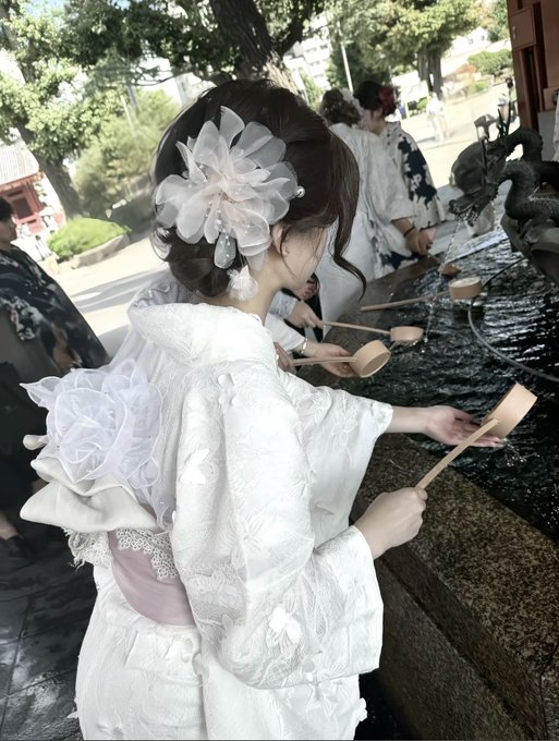 A woman in a traditional white kimono with an elaborate floral hair ornament participates in a purification ritual using a wooden ladle at a temizuya.