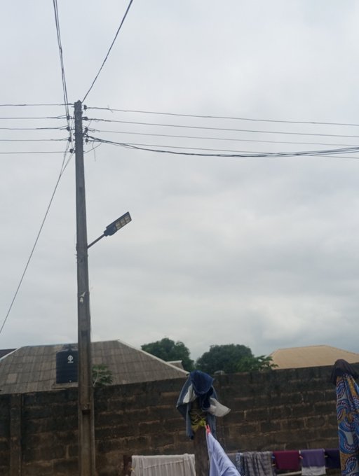 A street scene featuring a utility pole with a tangle of wires and a street lamp, set against a cloudy sky above a walled courtyard with laundry hanging out to dry.