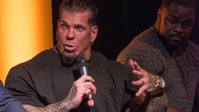 A tattooed muscular man with a goatee speaks into a microphone during what appears to be a panel discussion, while a man in a gray shirt stands behind him.