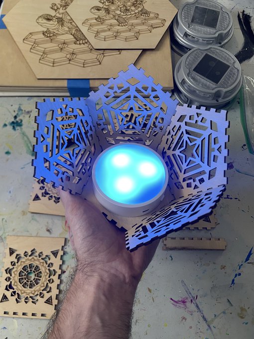 A hand holding a hexagonal wooden craft with LED lights inside, surrounded by other wooden crafts and solar lights on a workbench.