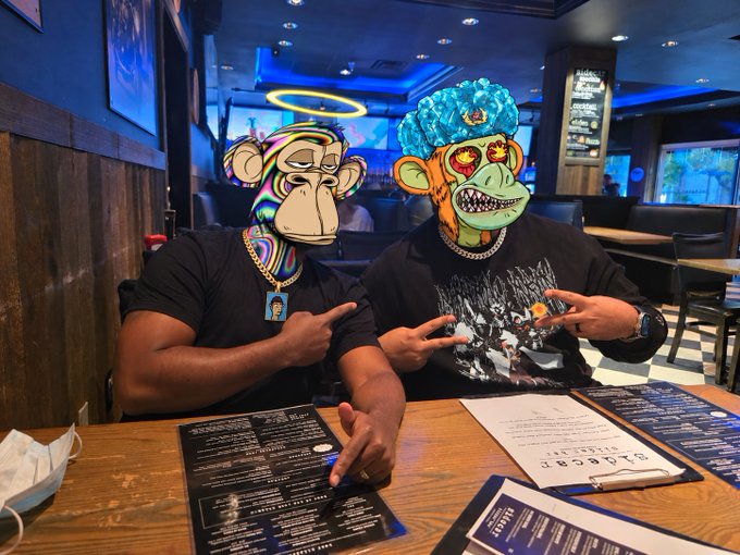 Two men sit at a restaurant table with menus; their faces are covered by Bored Ape Yacht Club NFT images, and they make hand gestures toward the camera.