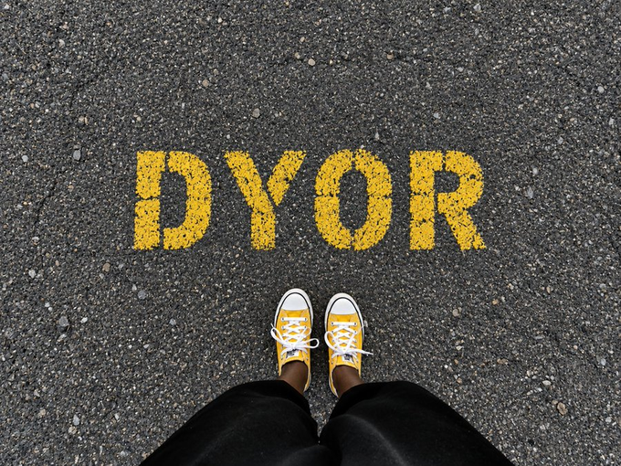 A first-person view shows someone wearing yellow Converse sneakers and black pants standing on asphalt with the word 'DYOR' (Do Your Own Research) painted in yellow letters on the ground in front of them.