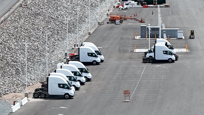 An aerial view of an industrial parking lot with a row of white electric semi-truck cabs parked along a large rock embankment.