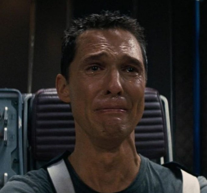 A close-up of a man with short dark hair crying, wearing a dark shirt with white straps over his shoulders.