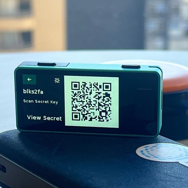 The image shows a small green device with a QR code displayed on its screen. The device appears to be a tablet computer, possibly an iPad or similar device. The QR code reads "View Secret Key", which suggests that the user might need to scan their 2FA account for verification purposes. The post also mentions the importance of hardware security and the need to export 2FA information as a QR code or readable secret key.