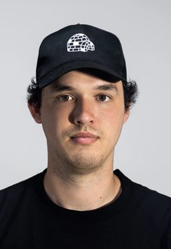 A man with dark hair and a slight stubble beard wears a black cap featuring a white outlined igloo logo, paired with a black t-shirt against a plain white background. The image accompanies news about Luca Netz, CEO of Pudgy Penguins, discussing his vision to IPO while integrating token and NFT holders.