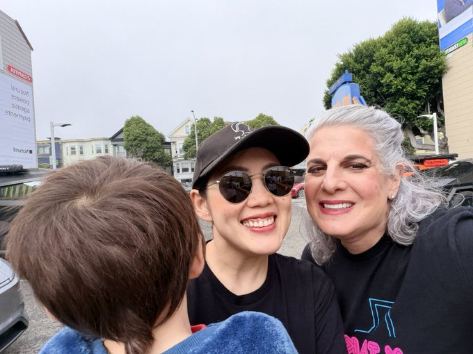 A woman wearing a black cap and sunglasses smiles alongside an older woman with gray hair while taking a selfie with a young boy in an outdoor urban setting.