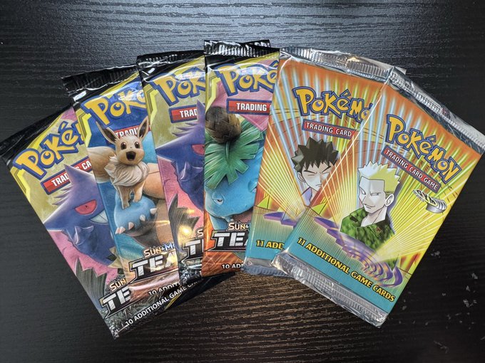 Five Pokémon Trading Card Game booster packs are fanned out on a wooden surface, including three from the "Team TE" Supreme set featuring Ivysaur, Eevee, and Gengar, alongside two Prism Star "11 Additional Game Cards" packs. The packs appear ready to be opened and added to the PokeHEX Time Warp Mystery Boxes mentioned in the post.