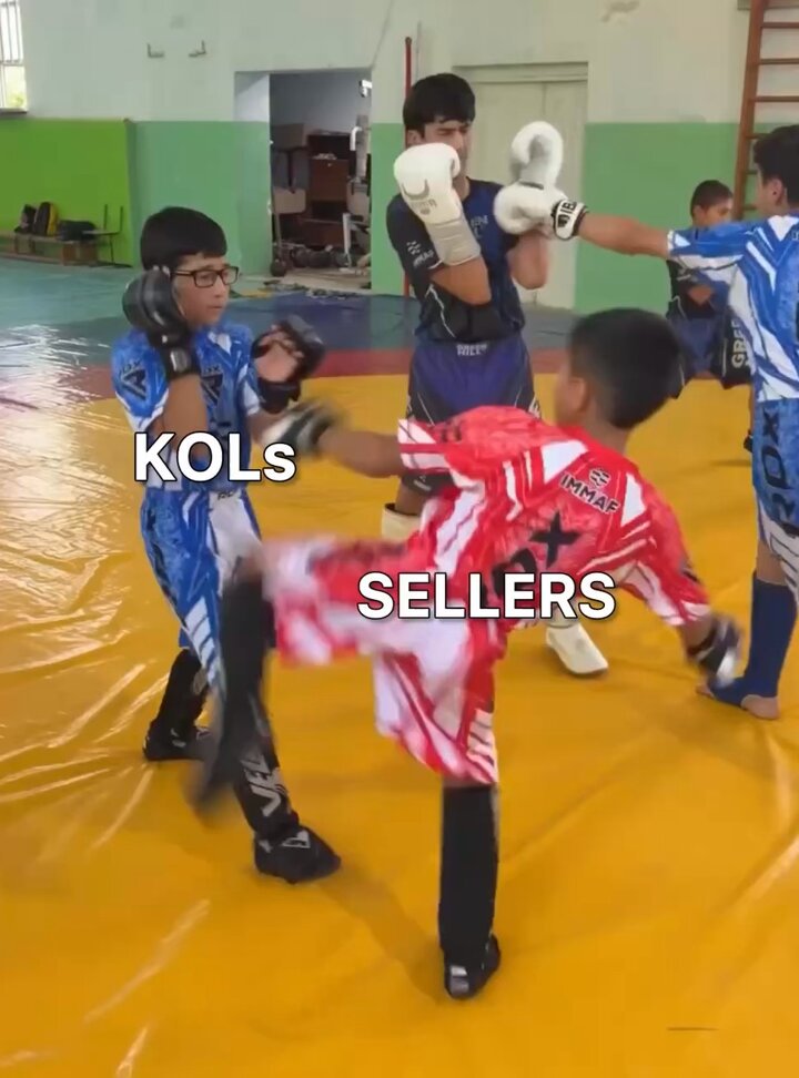 The image shows a group of young boys wearing boxing gloves, practicing their moves on an indoor gym floor. There are at least five children actively engaged in the activity, with one boy in the center of the action, kicking another boy who is wearing white gloves. The other kids appear to be learning from him or waiting for their turn to practice.