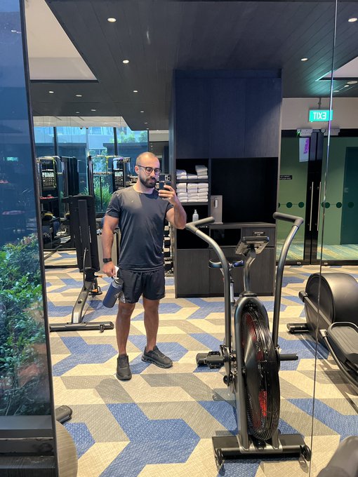 A man wearing a grey t-shirt and shorts takes a mirror selfie with his phone while holding a blue water bottle in a gym, standing next to exercise equipment.