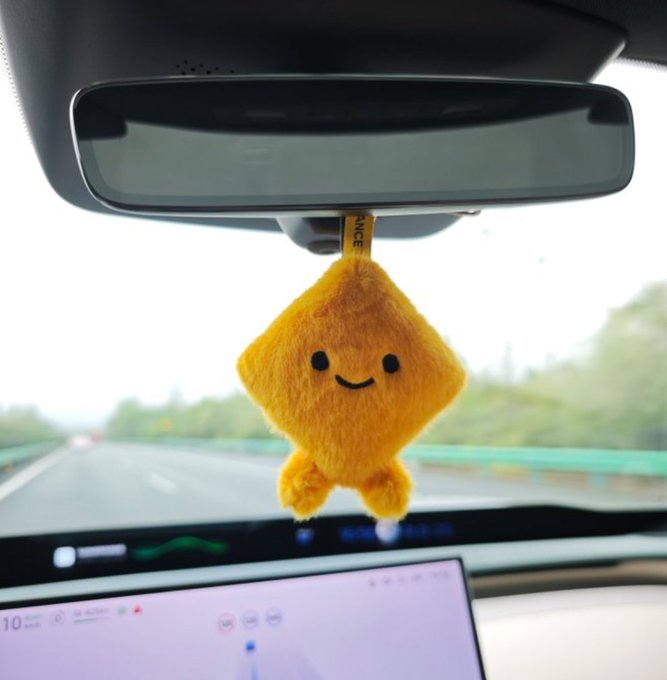 A small yellow plush star toy with a smiling face hanging from the rear-view mirror of a car driving on a highway, with the vehicle's infotainment screen visible below.