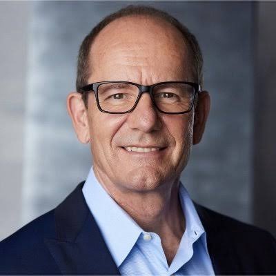 A professional headshot of a smiling, balding older man wearing black-framed glasses and a blue shirt under a dark blazer.