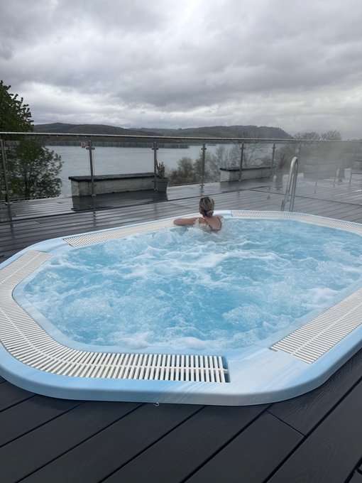 A person relaxes in a bubbling, light blue outdoor hot tub situated on a wooden deck, with a cloudy sky and a body of water visible in the background.