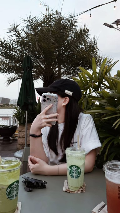 A woman wearing a black cap takes a mirror selfie at Starbucks with three iced drinks on the table, one prominently displaying the green Starbucks logo and "STARBUCKS" text. The relaxed weekend café setting complements the post's cheerful Saturday morning message about staying motivated with green candles and growing profits.