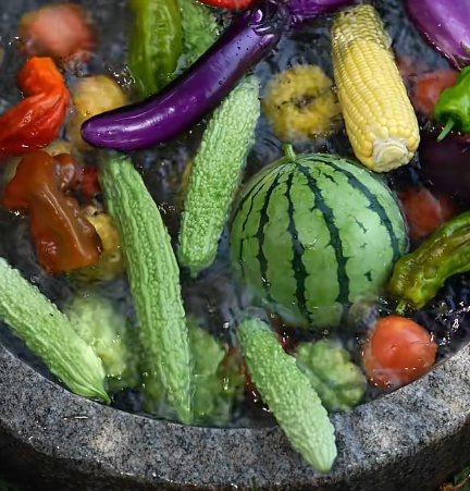 An overhead view of a stone bowl filled with water and an assortment of vegetables including a small watermelon, bitter melon, eggplant, baby corn, and peppers.