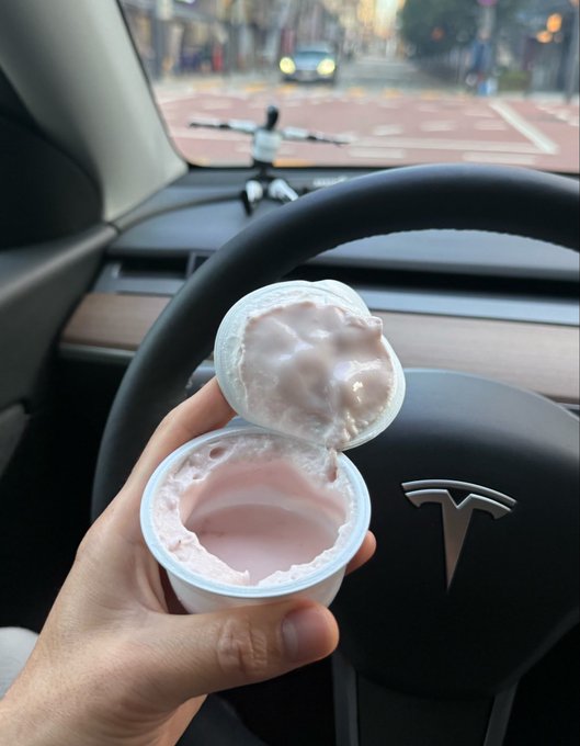 A hand holds an open cup of pink yogurt or dessert in front of a Tesla steering wheel featuring the Tesla 'T' logo, with a view of a city street through the windshield.