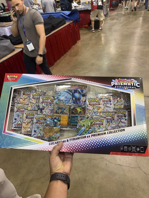 A first-person view at a convention showing a hand holding a Pokémon Trading Card Game 'Prismatic Evolutions' Lucario ex & Tyranitar ex Premium Collection box, with vendor tables and attendees visible in the background.