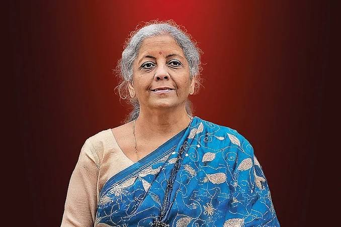 A woman with grey hair wearing a blue floral sari and beige blouse stands against a deep red background, with a small bindi on her forehead and a black beaded necklace visible. The image does not depict or relate to the post's content about India's Long-Term Capital Gains (LTCG) tax figures, as no text, numbers, or identifiable figures from the parliamentary reply appear in the photograph.