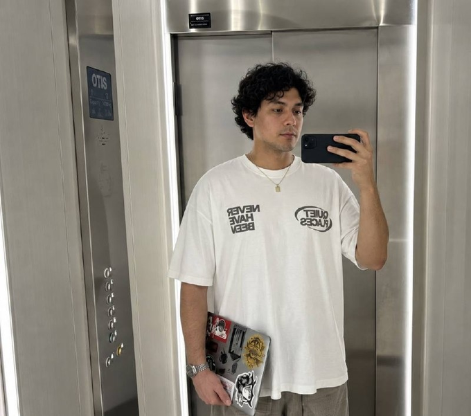 A young man with curly dark hair taking a mirror selfie in an OTIS elevator while wearing a white t-shirt with mirrored 'NEVER HAVE I EVER' and 'QUIET ZOOM' text graphics.