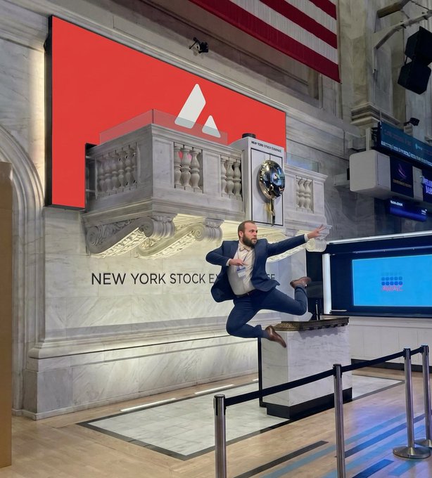 A man in a blue suit joyfully leaps off a marble pedestal at the New York Stock Exchange, where the iconic red NYSE sign and American flag hang in the background. The image captures the spontaneous excitement suggested by the caption "just saw these" — a moment of celebration or surprise on the trading floor.