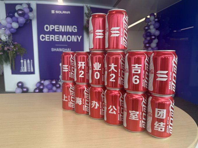 Stacks of red Coca-Cola cans featuring custom Chinese characters spell out "开业大吉" (wishing prosperity for your new business) and "上海办公室团结" (Shanghai office unity), displayed against a backdrop reading "SOLANA OPENING CEREMONY SHANGHAI," celebrating Solana's new Shanghai office launch.