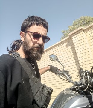 A man with a dark beard and sunglasses stands outdoors next to a motorcycle, wearing a black shirt and a shoulder bag, with a brick wall and trees in the background.