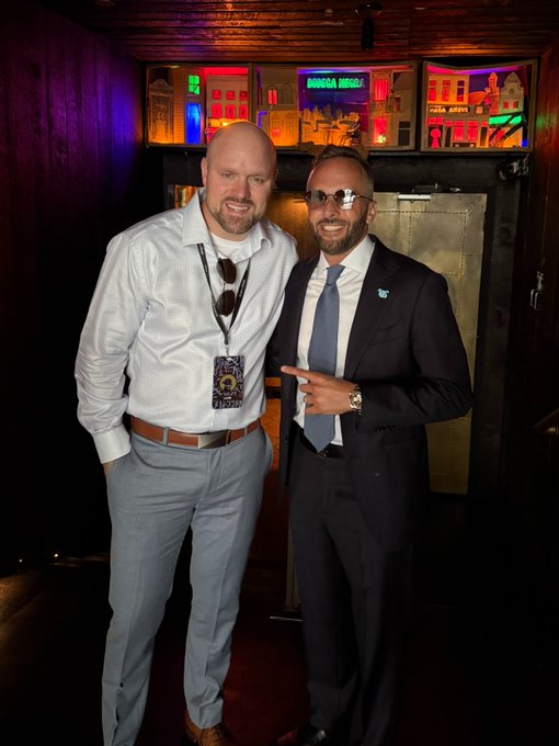 Two men posing together indoors; one is bald wearing a white shirt with a lanyard, and the other wears a dark suit, blue tie, and sunglasses, pointing at his companion.