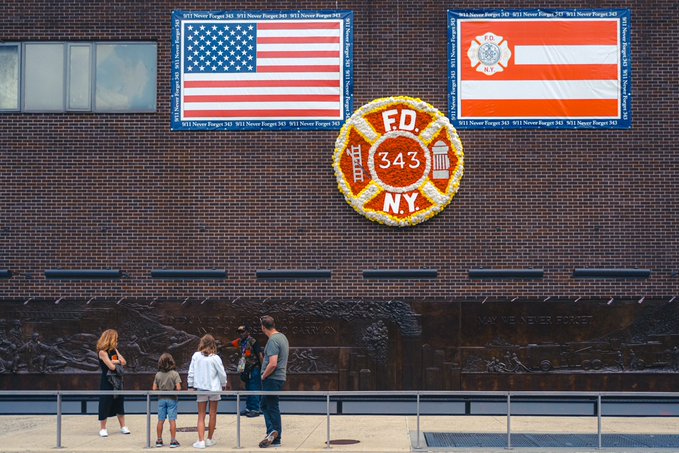 A family stands viewing a memorial display on a brick wall featuring an American flag, a FDNY 343 emblem made of flowers, and another flag with '911 None Forget 343' text around the borders.