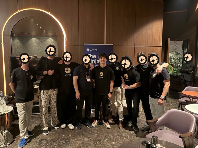 The image shows a group of people posing for a photo, all wearing black t-shirts with white text that says "ASTER" on them. The group consists of at least eight individuals standing close together, some holding drinks or cups. In the background, there is an archway and a sign that reads "THE JOURNAL".