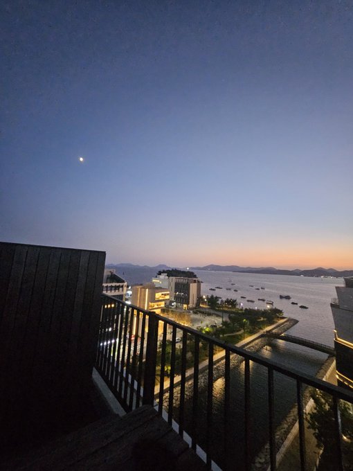A scenic view from a balcony at dusk overlooking a coastal city with a large cruise ship docked at a pier, mountains in the distance, and a crescent moon visible in the sky.