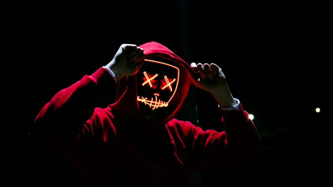 A person wearing a red hoodie with an illuminated LED mask featuring X-eyes and a stitched smile, pulling the hood up in a dark setting.