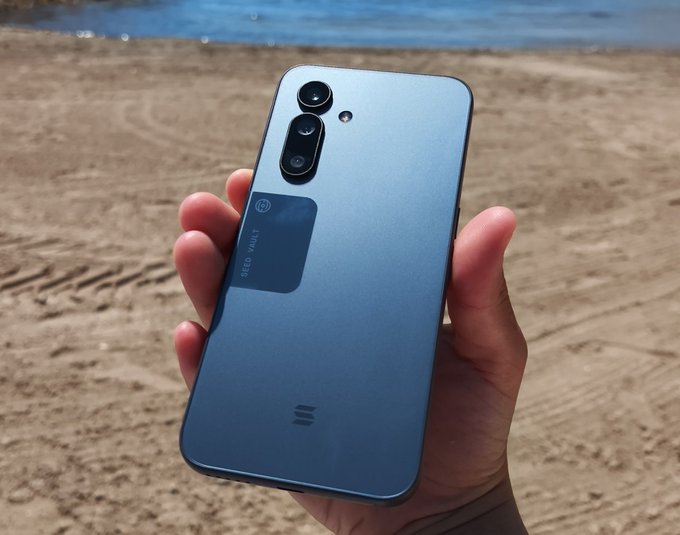 A hand holding a blue smartphone (appearing to be a Meizu 21) at the sandy beach with the ocean in the background, showing the rear camera module and "MELON" branding sticker.