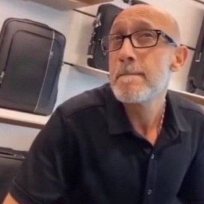 A bald man with glasses and a white beard wearing a black polo shirt looks confused or taken aback, seated indoors with black cases visible on shelves behind him, capturing the puzzled "Sorry what?" reaction.