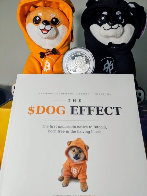 Two plush dog toys wearing bitcoin-themed hoodies (orange and black) sit behind a silver Bitcoin coin and a book titled 'The $DOG EFFECT - A Cryptolution Research Handbook, 2026 Edition.'