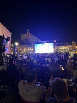 The image shows an outdoor event where people have gathered around a large screen displaying a soccer game, likely the World Cup. The crowd consists of many individuals sitting at tables or standing, all watching the game intently. There are also several chairs scattered throughout the scene. In addition to the spectators and their drinks, there is a bottle visible in the image. The event appears to be well-attended with people enjoying the atmosphere and cheering for their favorite teams.
