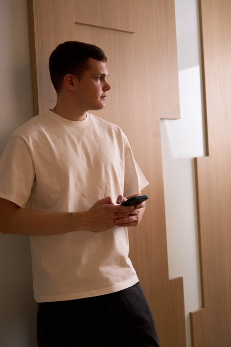 A young man in a cream-colored t-shirt stands leaning against a wooden panel, holding a phone and gazing off to the side, projecting a thoughtful, professional demeanor fitting for a 24-year-old podcaster seeking new connections.