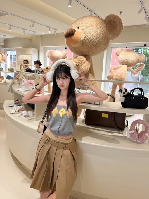 A young woman wearing a fuzzy bear ear headband, a grey argyle top, and a pleated tan skirt poses inside a store filled with large teddy bears and handbags.