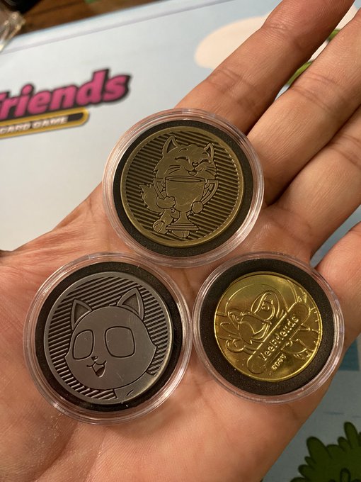 A hand holds three VeeFriends collectible coins in protective cases, featuring designs of a cat with a trophy, a cat face, and a cartoon character with text reading "A SQUIRREL'S SAGA" and "AT 09 12." The coins are displayed alongside VeeFriends branded materials in the background, matching the post's reference to "Vintage @veefriends coins."