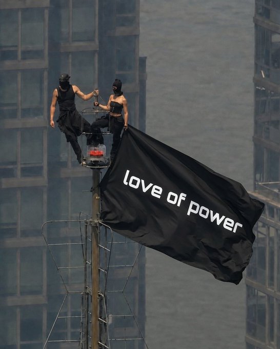 Two people dressed in black ninja-like attire standing on a tall pole, unfurling a large black flag that reads 'love of power' against an urban skyline.